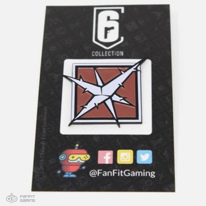 May include: A black and white enamel pin featuring a red square with a white starburst inside. The pin is on a black card with the text "Rainbow Six Collection" and a logo for Ubisoft Entertainment. The card also includes a social media handle for FanFitGaming.