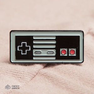 May include: A rectangular enamel pin featuring a classic video game controller design. The pin has a black background with a gray center section and white details. Two red buttons are on the right side, and a directional pad is on the left.