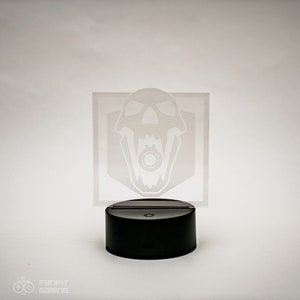 May include: A white acrylic night light featuring a skull design. The skull has a ring in its mouth and is enclosed within a hexagon shape. The night light sits on a black base with a button on the front.