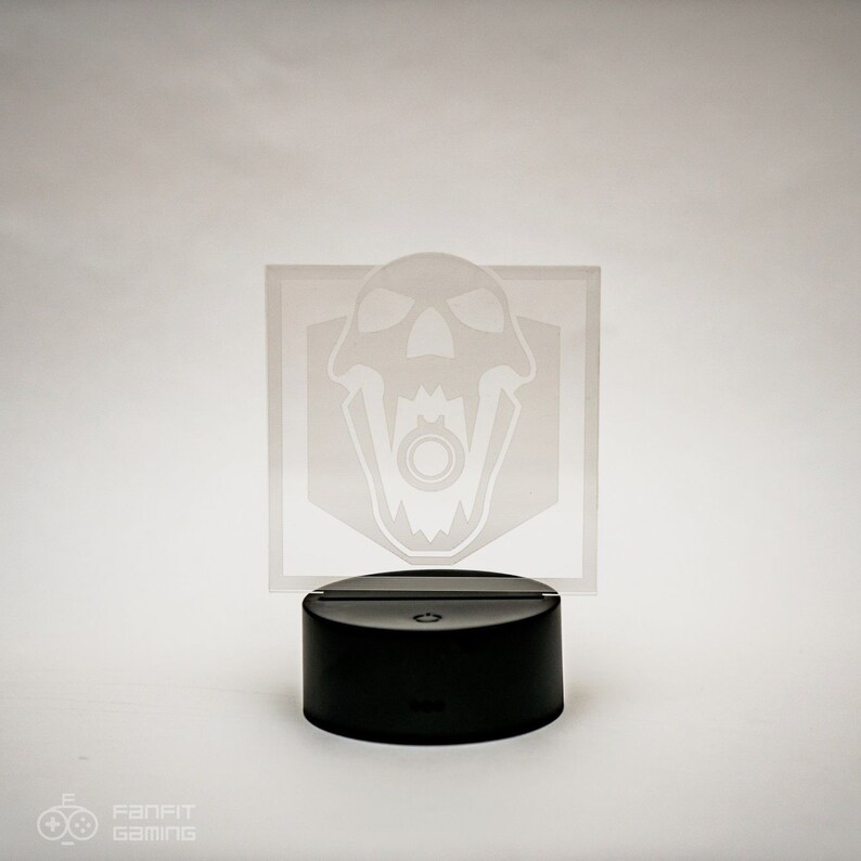 May include: A white acrylic night light featuring a skull design. The skull has a ring in its mouth and is enclosed within a hexagon shape. The night light sits on a black base with a button on the front.