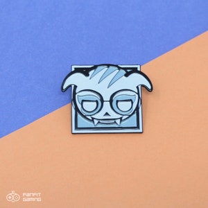 May include: A white and black enamel pin featuring a cartoon character with a skull-like face wearing glasses. The character is in a square frame with a black outline.