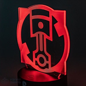 May include: Red acrylic lamp with a circular design featuring a stylized piston inside a circle. The lamp is on a black base.
