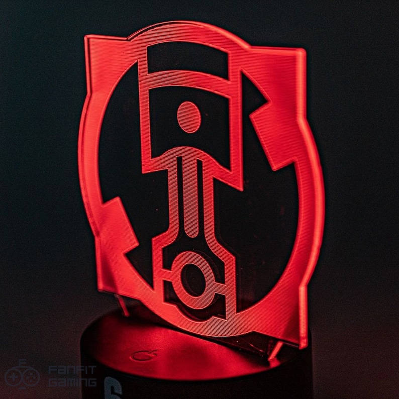 May include: Red acrylic lamp with a circular design featuring a stylized piston inside a circle. The lamp is on a black base.