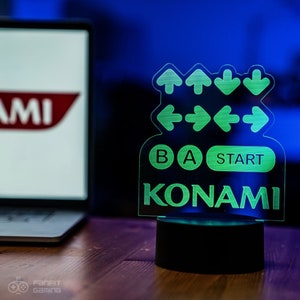 Może przedstawiać: Zielony podświetlany znak z kodem Konami, zawierający strzałki, litery B, A i słowo "START", a także słowo "KONAMI" w zielonym kolorze.