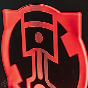 May include: A red 3D printed logo with a piston design. The piston is outlined in black and has a red circle in the center. The logo is surrounded by a red circle.