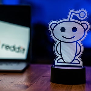 May include: A white Reddit alien logo night light with a black base. The logo is a stylized alien with a smile and antennas.