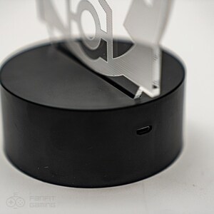 May include: A black circular base with a USB charging port on the side. A clear acrylic figure sits on top of the base.