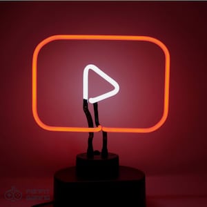 May include: Neon sign in the shape of the YouTube logo. The sign features a bright orange rectangular border with rounded corners and a white play button in the center. The sign is mounted on a black base.