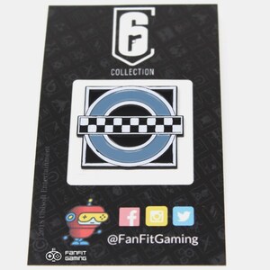 May include: A black and white enamel pin featuring a circular design with a checkered stripe across the center. The pin is surrounded by a blue border and a black square. The text "Rainbow Six Collection" is printed above the pin.
