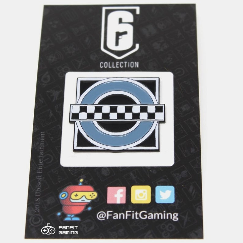 May include: A black and white enamel pin featuring a circular design with a checkered stripe across the center. The pin is surrounded by a blue border and a black square. The text "Rainbow Six Collection" is printed above the pin.