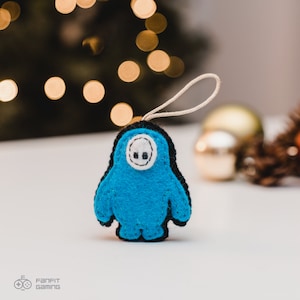 Puede incluir: Un adorno de fieltro azul con forma de personaje del juego Fall Guys. El adorno tiene una cara blanca con dos ojos negros y un contorno negro.