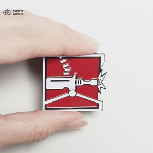 May include: A red and white enamel pin featuring a stylized image of a machine gun with a red background and a white outline. The pin is held in a hand.