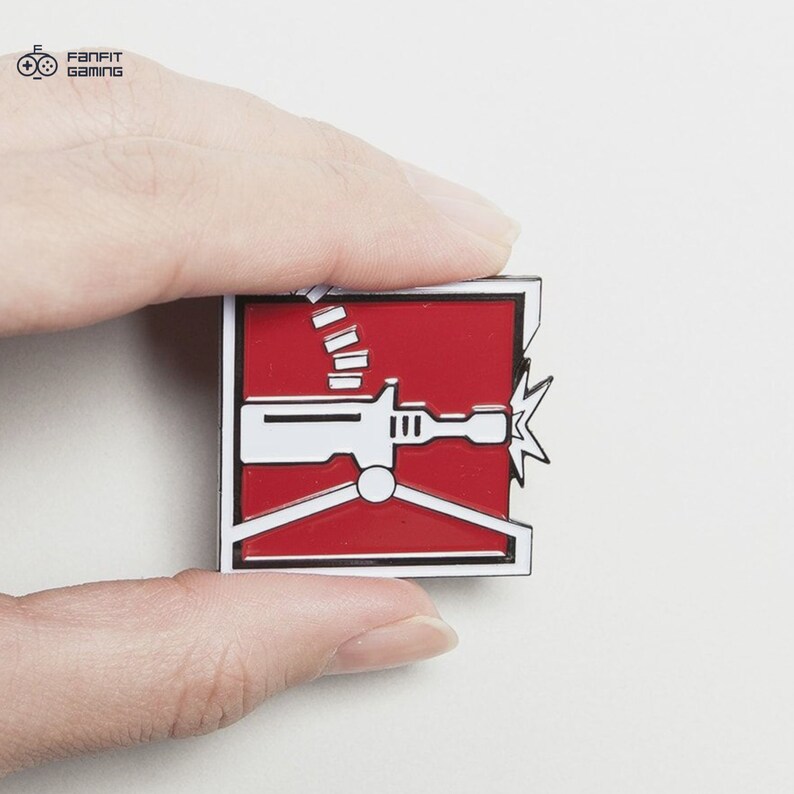 May include: A red and white enamel pin featuring a stylized image of a machine gun with a red background and a white outline. The pin is held in a hand.
