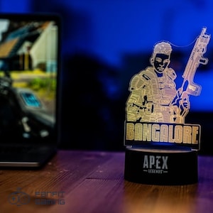 Op de afbeelding: Een 3D-geprint LED-nachtlampje met Bangalore, een personage uit de videogame Apex Legends. Het lampje is aan en toont een gele omtrek van Bangalore die een geweer vasthoudt. De basis van het lampje is zwart en heeft de tekst "Apex Legends" in wit.