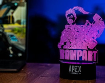 May include: A pink and purple 3D printed lamp with a silhouette of a video game character holding a weapon. The lamp base is black and has the text "Rampart" and "Apex Legends" printed on it.
