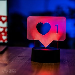 May include: A red and white illuminated acrylic light-up sign shaped like a social media like button with a heart and the number 1. The sign is on a black base.