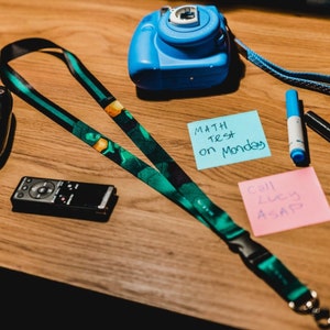 May include: A wooden desk with a black video game controller, a blue camera, a green and black lanyard, a blue and black marker, a black marker, a pink sticky note with the text "Call Lucy ASAP", and a blue sticky note with the text "MATH TEST on Monday".