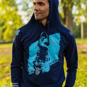 May include: A navy blue hooded sweatshirt with a graphic of a character from the video game Rainbow Six Siege. The character is wearing a blue and white tactical suit and is holding a gun. The text "FROST" is printed on the sweatshirt.