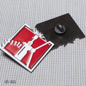 May include: A red and white enamel pin featuring a stylized image of a drill with a red background and a white outline. The pin is attached to a black metal backing with a pin and clutch.