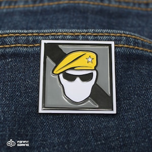 Puede incluir: Un pin de esmalte blanco y negro con fondo gris. El pin presenta una figura blanca enmascarada que lleva una boina amarilla con una estrella blanca. La figura lleva gafas de sol y tiene una línea diagonal negra detrás.