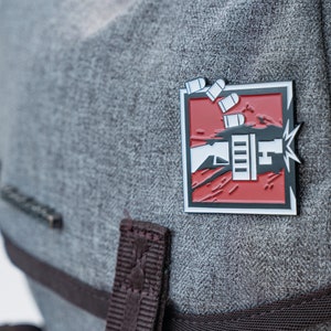 Puede incluir: Un pin de esmalte rojo y negro con una imagen estilizada de una mano sosteniendo una pistola con balas que salen del cañón. El pin está sobre un fondo de tela gris.