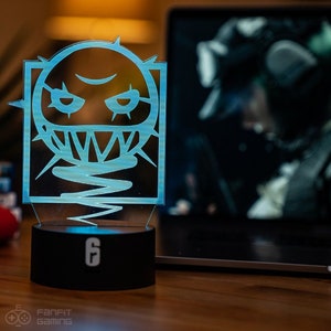 May include: A blue and white light-up lamp shaped like the Rainbow Six Siege operator,  "Lesion". The lamp has a black base with the number "6" on it.