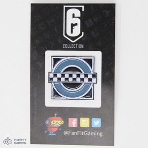 May include: A black and white enamel pin featuring a checkered pattern and a blue circle with a white outline. The pin is on a black card with the text "Rainbow Six Collection" and a logo for the game.