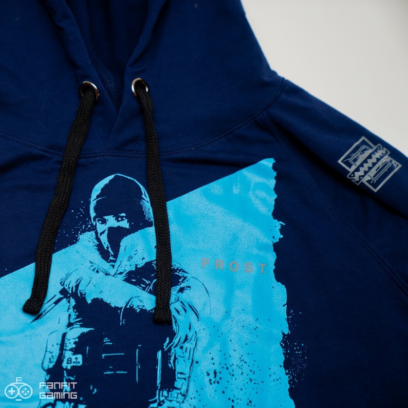 May include: A navy blue hoodie with a graphic of a character from the video game "Rainbow Six Siege". The character is wearing a blue and white mask and is holding a gun. The text "Frost" is printed on the hoodie.