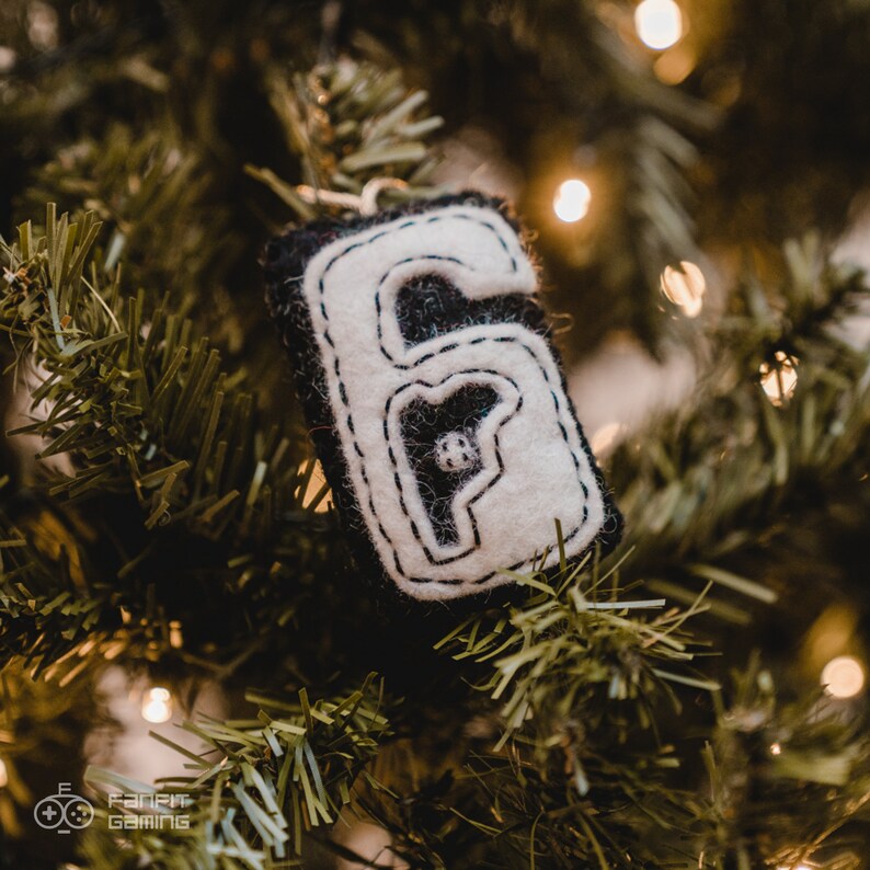 Puede incluir: Un adorno de fieltro blanco con forma de n&uacute;mero seis con costuras negras. El adorno est&aacute; colgado de un &aacute;rbol de Navidad verde.