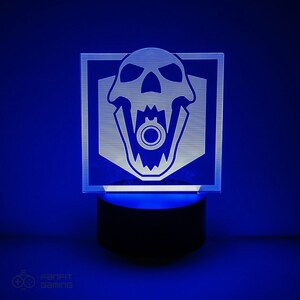 May include: A white acrylic night light featuring a skull design with a gun in its mouth. The light sits on a black base and is illuminated with blue light.
