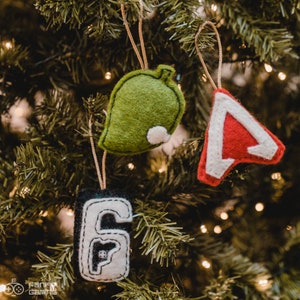 Puede incluir: Tres adornos navide&ntilde;os de fieltro colgados de un &aacute;rbol de Navidad verde. Los adornos son una hoja verde, un tri&aacute;ngulo rojo y blanco y un n&uacute;mero seis negro y blanco.
