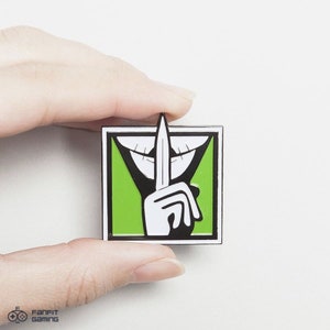 May include: A green and black enamel pin featuring a white hand holding a knife to its mouth, with a white smile behind the hand. The pin is a popular symbol from the video game Rainbow Six Siege.
