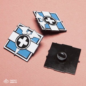 May include: Three enamel pins with a white cross on a blue and white geometric design. The pins have a black border and a black backing with a pin clasp. The pins are on a pink surface.