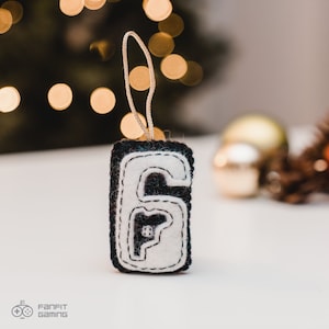 Puede incluir: Un adorno navide&ntilde;o de fieltro con forma de n&uacute;mero seis, con un interior de fieltro blanco y costuras negras. El adorno tiene un lazo de cuerda blanco para colgar.