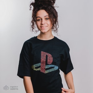 May include: A person wearing a black t-shirt with a colorful PlayStation logo made of video game titles. The logo is made up of words like "FIFA", "Fortnite", "Minecraft", and "Grand Theft Auto".