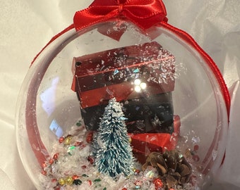 May include: A clear glass Christmas ornament with a red bow on top. Inside the ornament, there is a small artificial pine tree, red and black gift boxes, and red and gold glitter.
