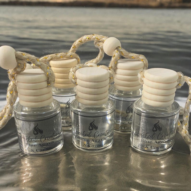 May include: Six small glass bottles with wooden tops and gold string hangers. The bottles are filled with a clear liquid and have labels that say "Sparked Creations Blue Agave & Cacao".
