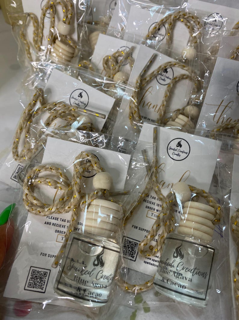 May include: A set of car air fresheners with a gold and white string. The air fresheners are in clear glass bottles with a wooden stopper and a label that says "Sparked Creations Blue Agava & Cacao".