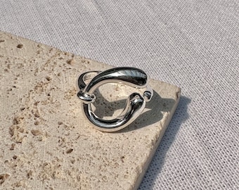 May include: A silver ring with a unique, abstract design. The ring is made of a thick, rounded band that forms a circle with a gap in the middle.