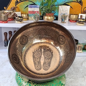 May include: A large, bronze singing bowl with detailed geometric patterns and a central design of two stylized footprints. The bowl is displayed on a green cushion. Other smaller bowls and spiritual items are visible in the background.