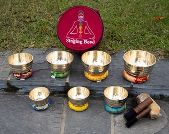 Puede incluir: Siete cuencos cantores de diferentes tamaños están dispuestos sobre una superficie de piedra gris. Cada cuenco descansa sobre un cojín de colores. Una bolsa roja con las palabras "Singing Bowl" está en el fondo. Dos mazos de madera están en el lado derecho de la imagen.