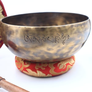 May include: A traditional Tibetan singing bowl with a hammered finish, resting on a red and gold cushion. The bowl has a  Tibetan inscription on the inside. A wooden striker with a black rubber tip is also included.