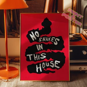 Op de afbeelding: Een rode poster met zwarte en witte tekst die "No Snakes In This House" zegt. De poster is in goud ingelijst en hangt aan een muur.