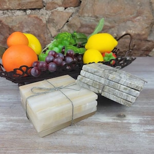 May include: Two sets of stone coasters tied with twine. One set is a light beige color, and the other is a gray and white marble pattern. A decorative metal tray filled with fruit, including oranges, lemons, and grapes, sits in the background.