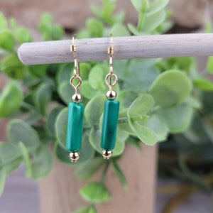 May include: A pair of dangle earrings featuring gold-colored hooks and small gold beads. Each earring has a cylindrical, vibrant green malachite stone. The earrings are displayed against a backdrop of green foliage.