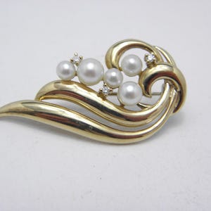 Puede incluir: Broche dorado con un diseño fluido y estilizado. El broche presenta varias perlas de imitación y pequeños acentos brillantes. La forma general recuerda a una ola o una hoja estilizada, con un cierre de alfiler en la parte posterior.