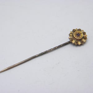 May include: A vintage gold-tone hat pin. The head of the pin has a floral design with a central purple stone. The pin has a twisted shaft and a sharp point. The pin is approximately 10 cm long.