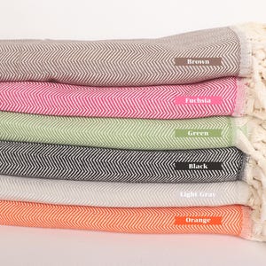 Peut inclure: Une pile de six serviettes en coton tissées de différentes couleurs, dont le marron, le fuchsia, le vert, le noir, le gris clair et l'orange. Chaque serviette a un motif en chevrons et des franges blanches.