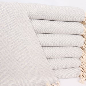 Puede incluir: Una pila de mantas de lana gris y blanca con patrón de espiga y flecos en los bordes. Las mantas están dobladas y apiladas sobre una superficie blanca.