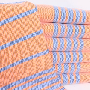 May include: A stack of orange and blue striped towels with white fringe. The towels are folded and stacked on a white surface.